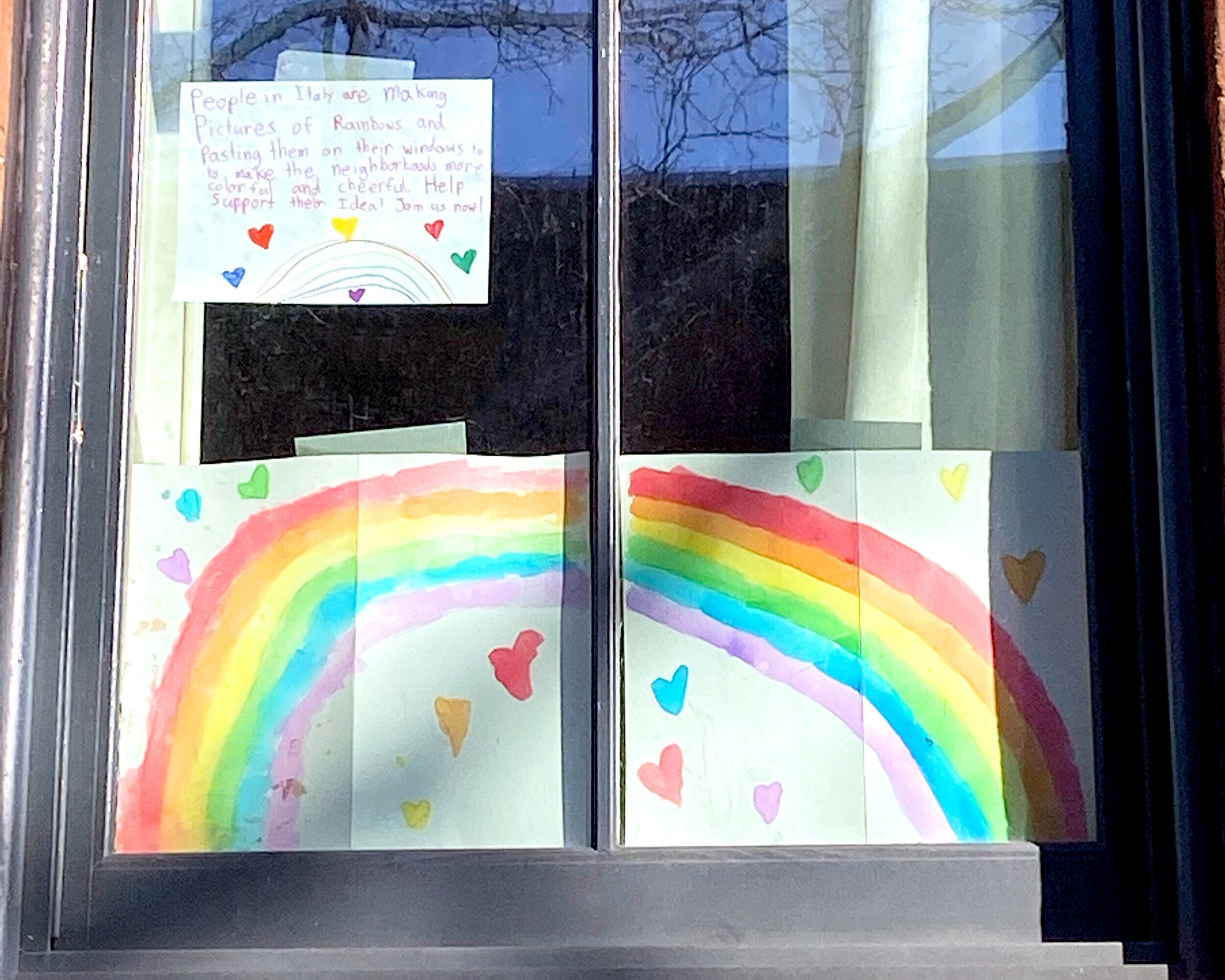 Child's hand-painted rainbow and heart artwork hanging in window with note that says "People in Italy are making pictures of rainbows and pasting them on their windows to make the neighborhoods more colorful and cheerful.