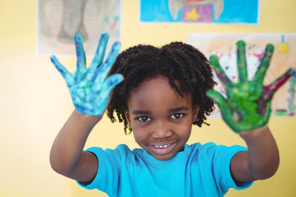 Smiling child in classroom holding up hands with blue and green paint on them