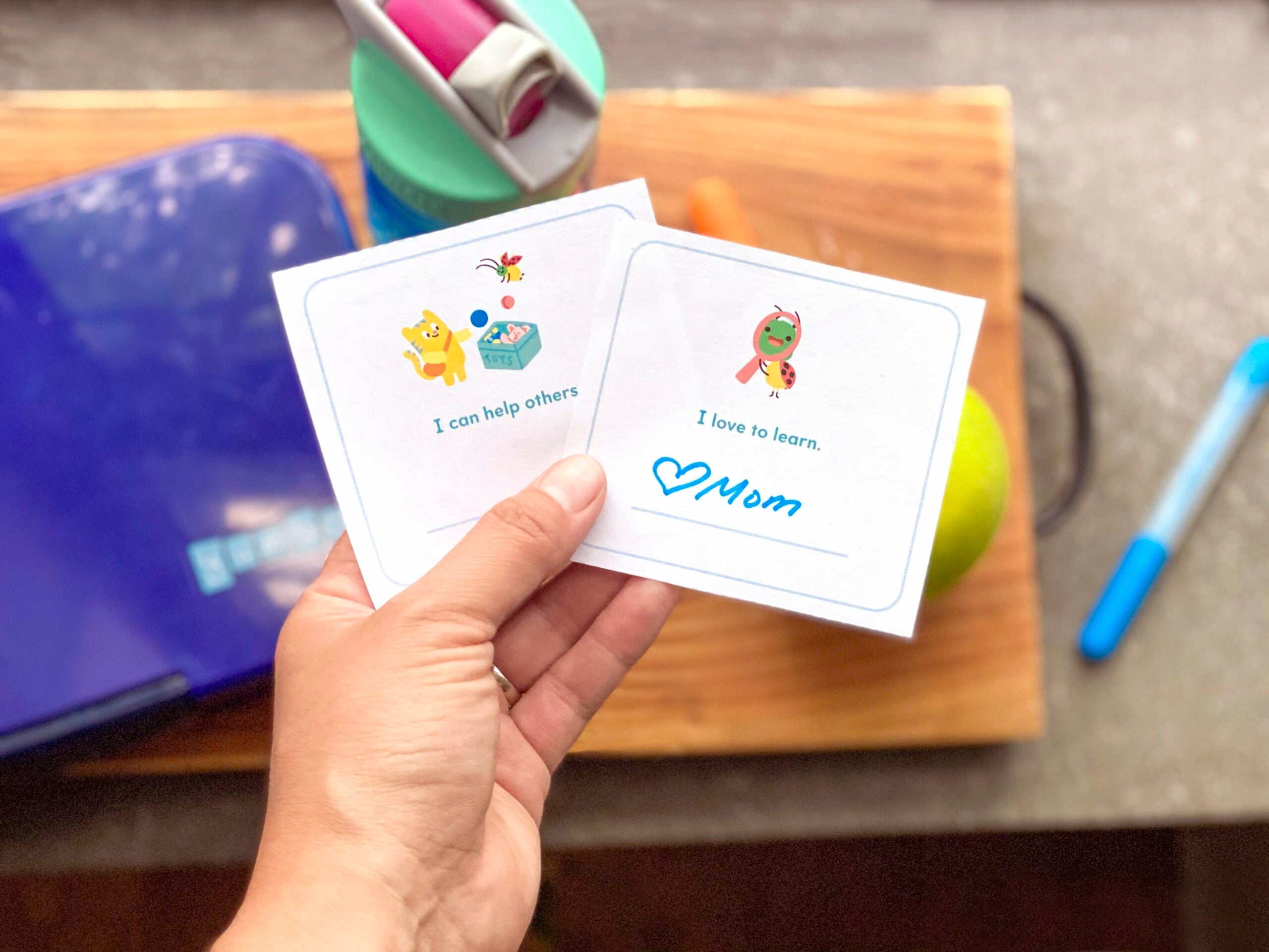 Free printable lunchbox notes held in a parent's hand over a lunchbox, apple, and water bottle