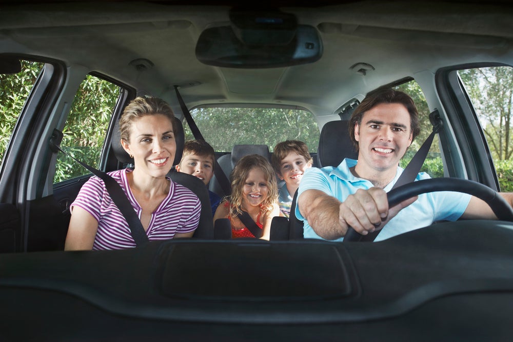 Family sitting in car wearing seatbelts, smiling at camera