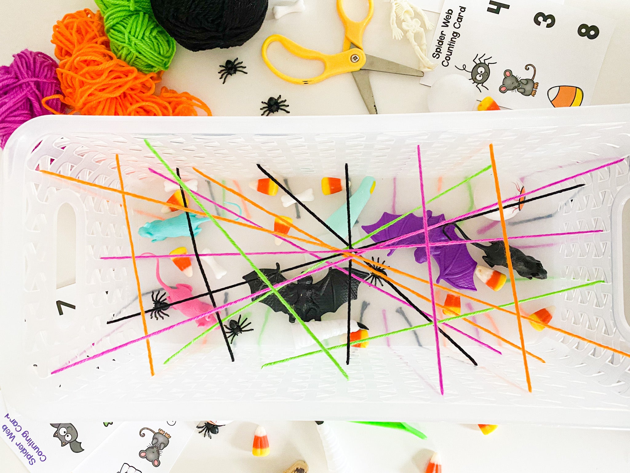 Spider web activity in progress; colored string through basket with candy and toys underneath, with printable and scissors beside it on a table