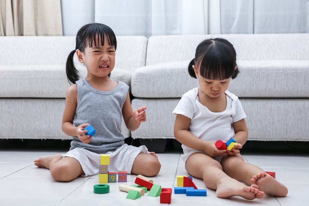 Girl crying while playing with blocks while her sister plays quietly next to her, unbothered