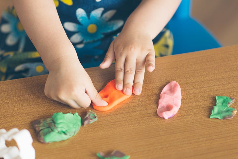 Child's hands stretching out orange play dough, with other play dough chunks around her in pink, green, and white