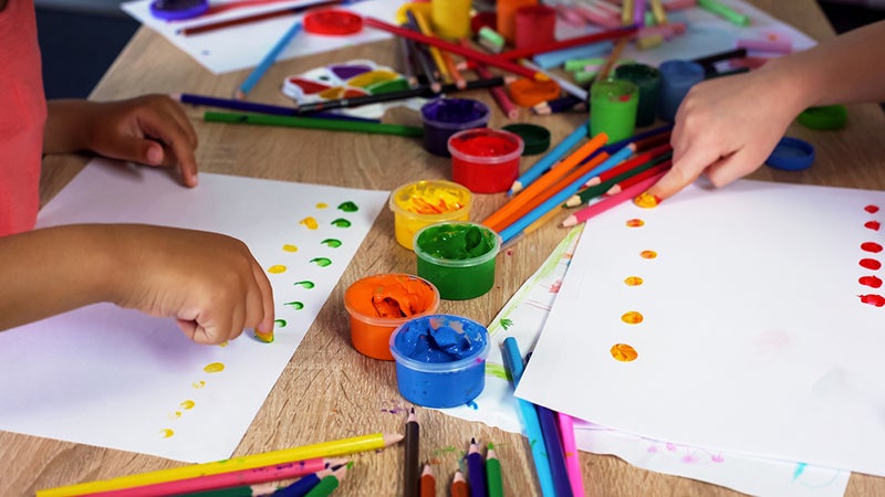 Children doing dot activities on paper, surrounded by paints