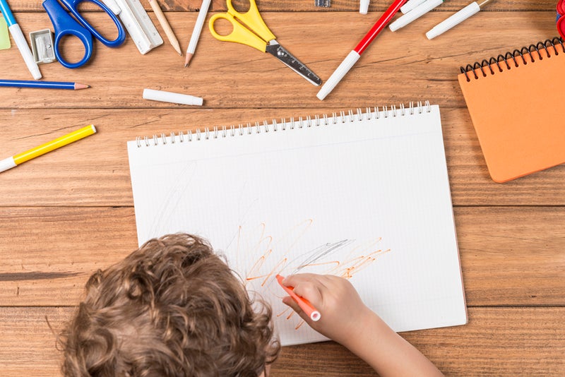 Child scribbling on a sketch pad, surrounded by scissors and markers