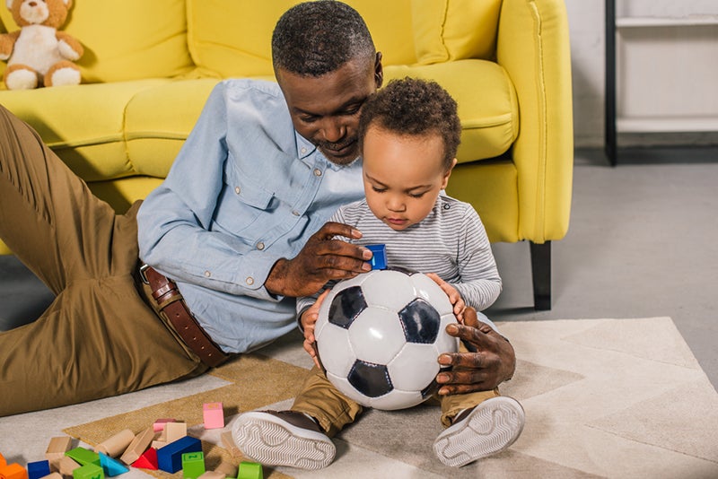 Grandparent playing with toddler inspecting blue block and black-and-white soccer ball on floor