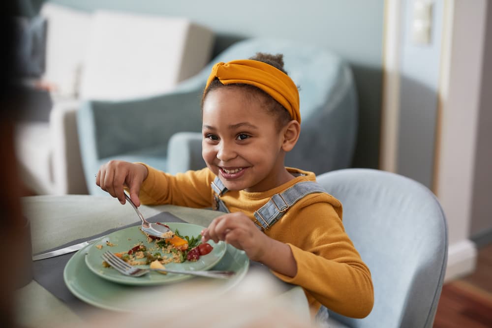 Toddler smiling while eating dinner with variety of healthy foods on plate
