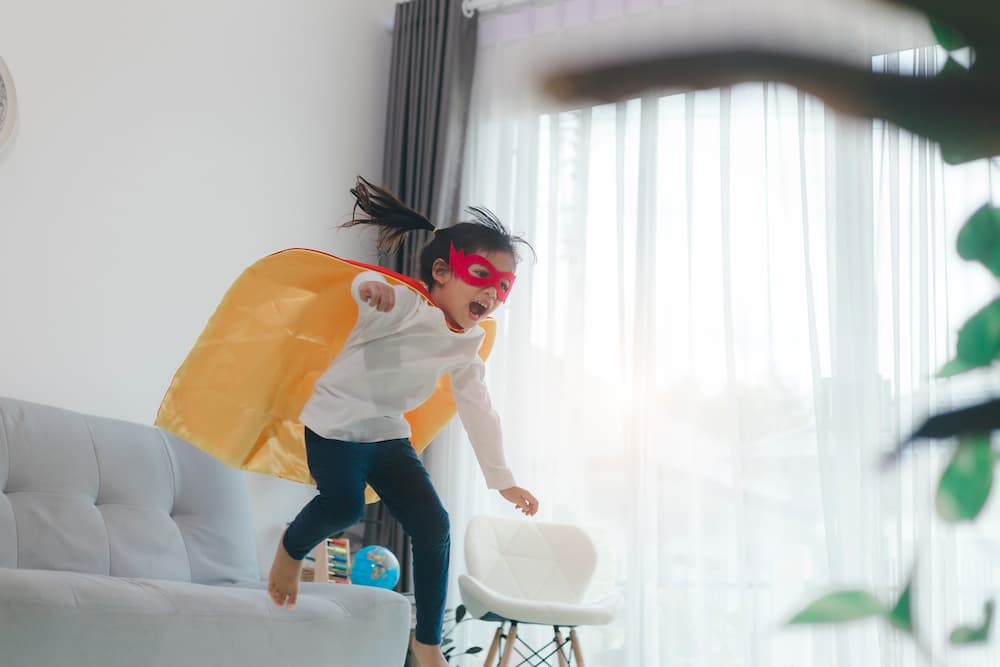 Girl in superhero costume jumping off couch indoors