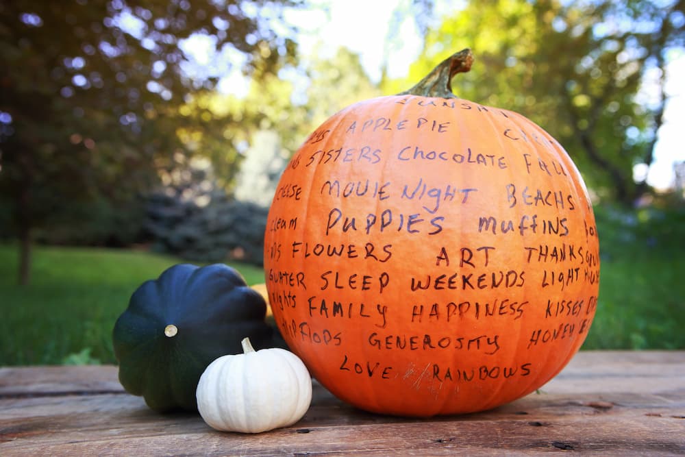 Thankful pumpkin covered in words describing what people are grateful for on table next to two other gourds