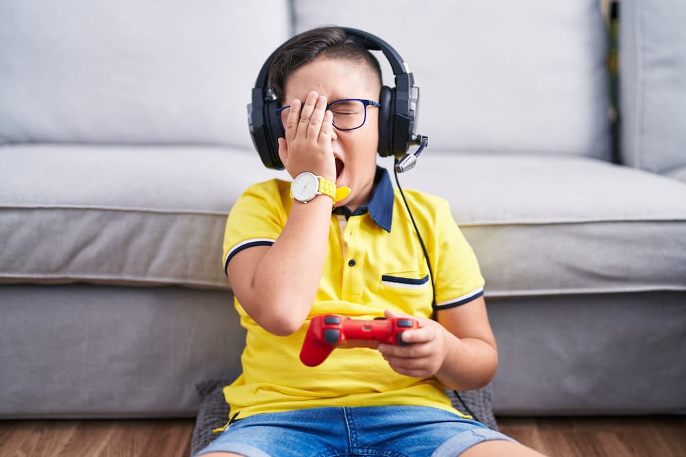 Child rubbing their eyes and yawning while sitting on floor holding a video game controller and wearing headphones